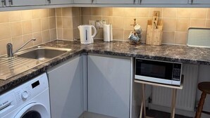 Cocina compacta con armarios blancos, lavadora integrada, microondas y cafetera.