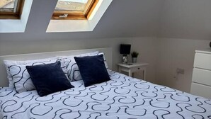 Un dormitorio con una cama, dos almohadas, una mesita de noche, una lámpara y un aparador. Hay tragaluces sobre la cama.