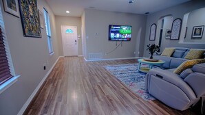 A living room with hardwood flooring, a gray sofa, a television mounted on the wall, and a door leading to another room.