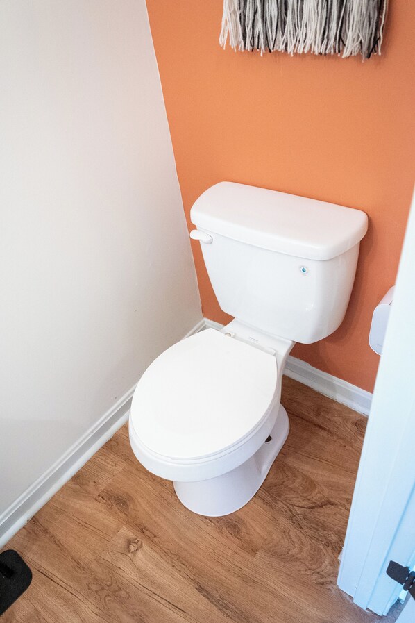 A white toilet with a closed lid, positioned against an orange wall, with a wooden floor and a white baseboard. - Cozy 3-bedroom house with Character in Waldorf (Waldorf)