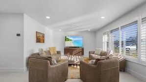 A modern living room with a beige sofa set, a flat-screen TV, and a large window with blinds.