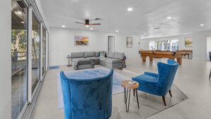 A spacious living area with a grey sectional sofa, blue armchairs, a pool table, and a ceiling fan.