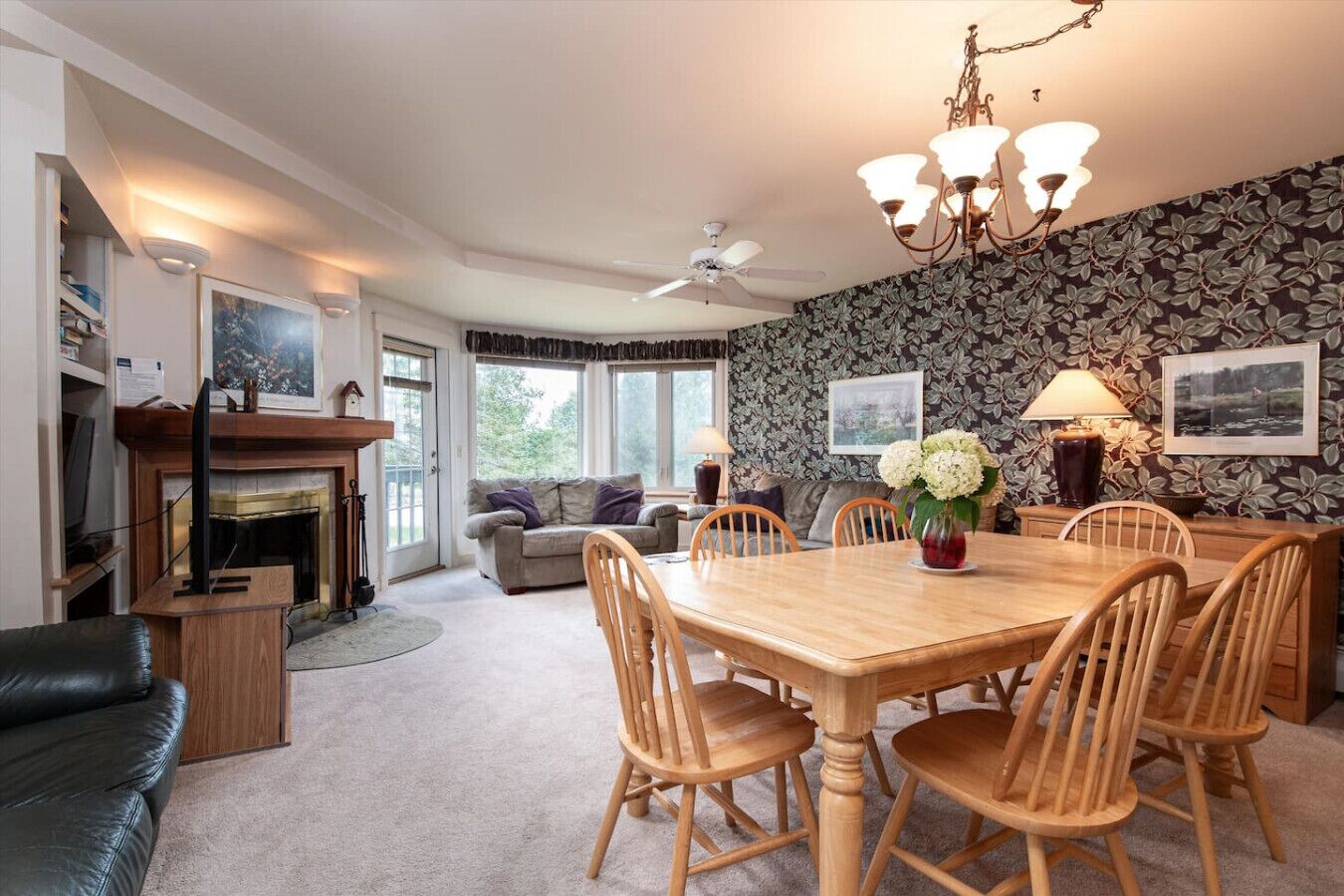 A cozy living room with a fireplace, a dining table with chairs, a sofa, and a ceiling fan.
