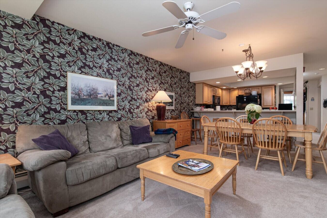 A living room with a sofa, coffee table, and a dining area with chairs and a table. There is a ceiling fan, a chandelier, and a kitchen in the background.
