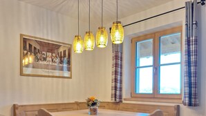 Un coin repas avec une table et des bancs en bois, des luminaires suspendus, une fenêtre avec des rideaux et un tableau encadré au mur.