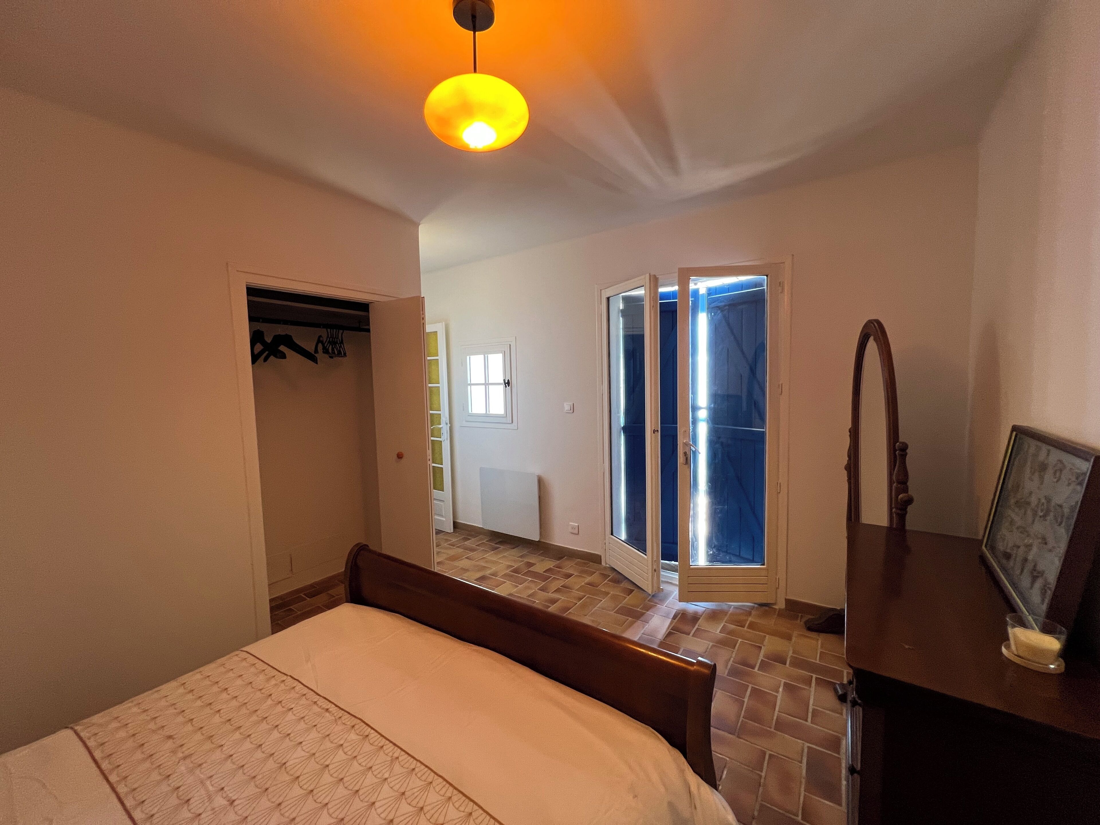Quarto com cama, cabeceira de madeira, mesa de cabeceira, espelho e porta para uma varanda.