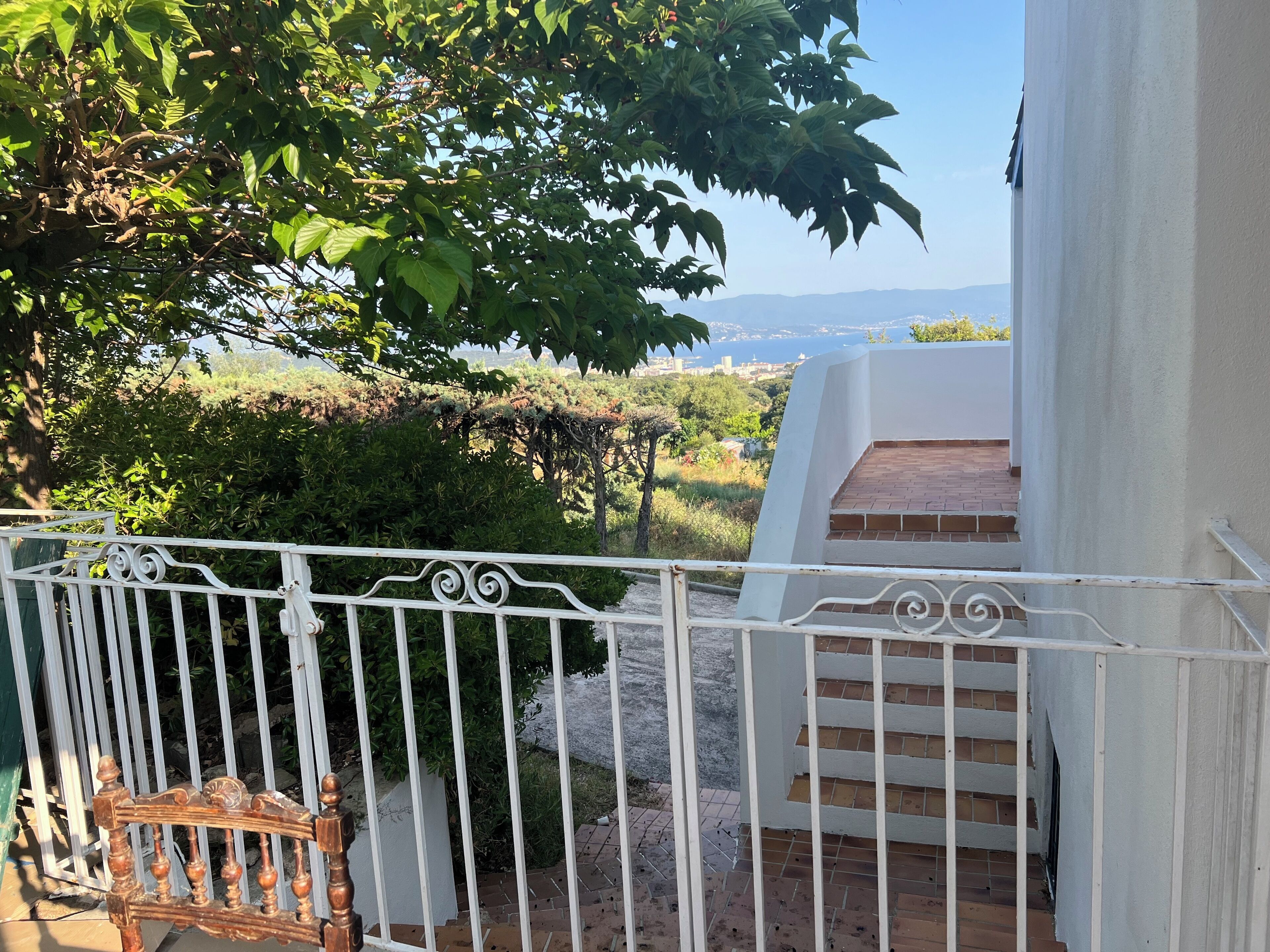 Ein Balkon mit weißer Brüstung, einer Holzbank und Blick auf einen angelegten Garten sowie eine entfernte Stadtlandschaft.