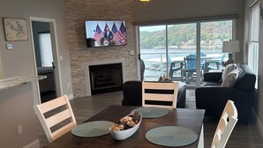 A dining area with a dark wooden table, white chairs, and a central bowl. A television mounted on the wall displays flags and a seated figure.