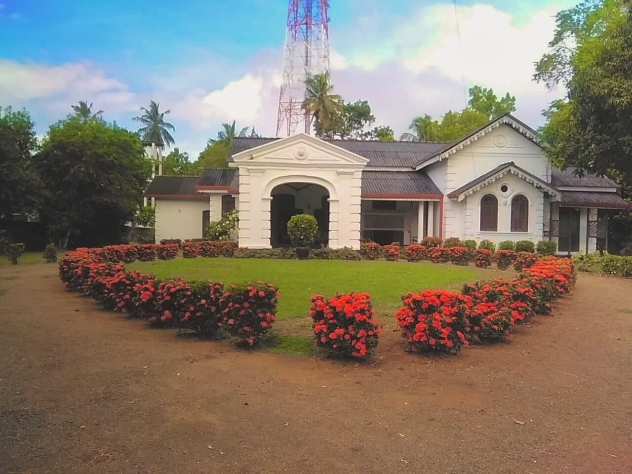 A building with a central archway, surrounded by a garden with red flowers, and a tall tower in the background.