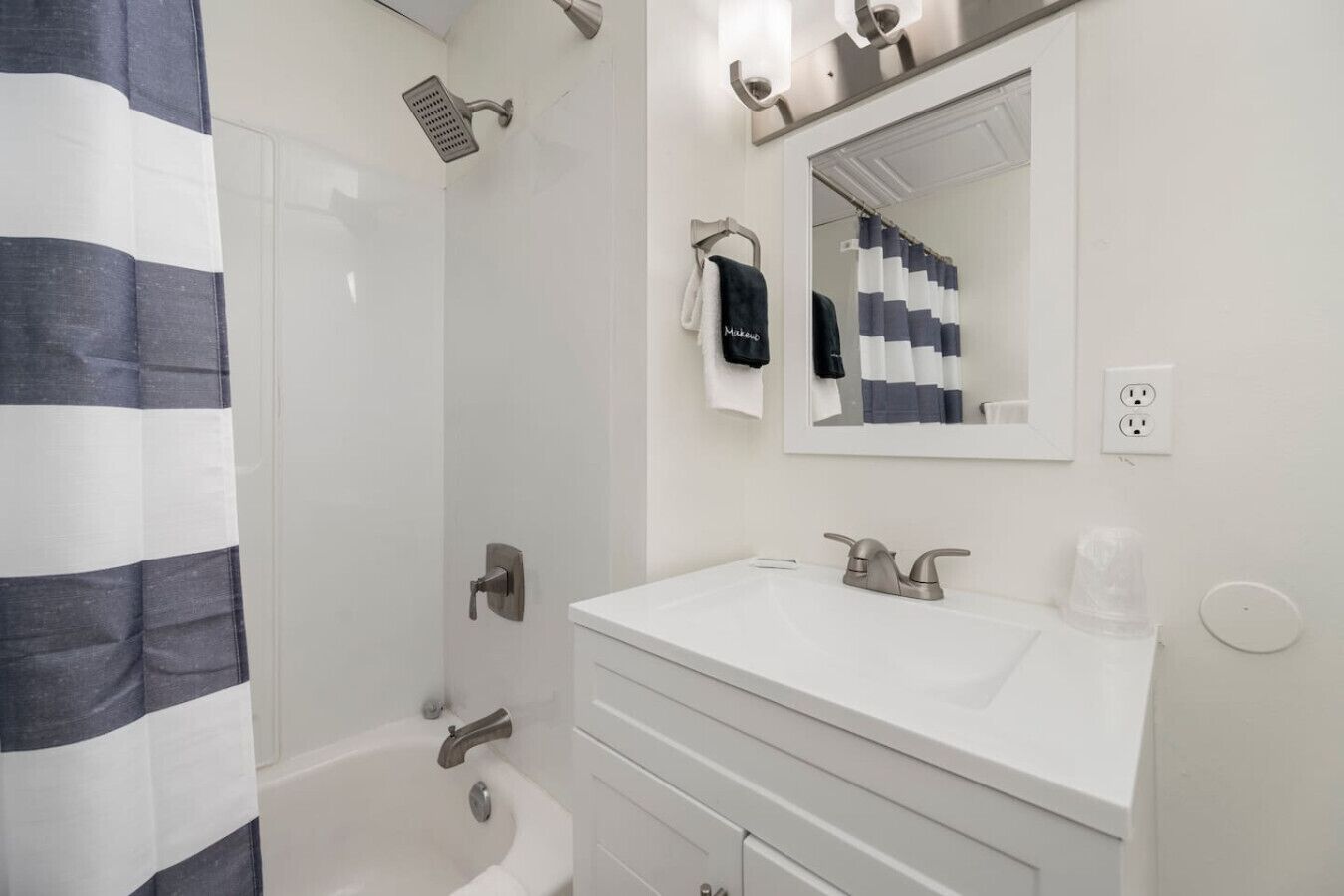 A modern bathroom with a white vanity, a large mirror, a shower with a handheld showerhead, and a towel rack with a black towel.