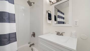 A modern bathroom with a white vanity, a large mirror, a shower with a handheld showerhead, and a towel rack with a black towel.