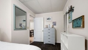 Una habitación de hotel con una cama, un aparador, un espejo, una puerta que da a un pasillo y una pared con un cartel enmarcado.