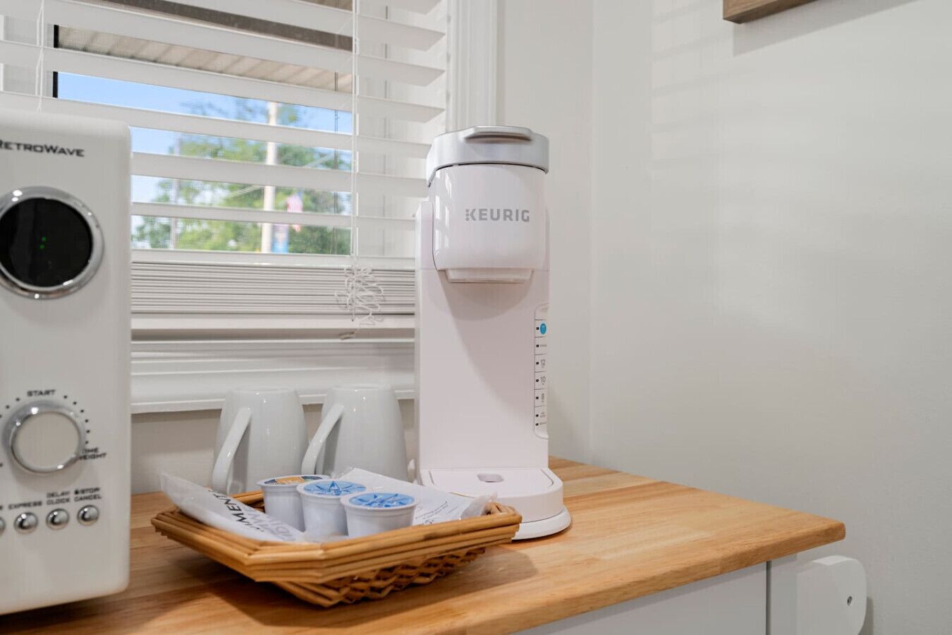 A coffee maker and a kettle on a wooden countertop near a window with blinds.