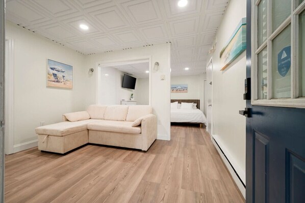 A room with a beige sectional sofa, a bed with white bedding, a flat-screen TV on the wall, and a wooden floor.