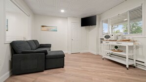 A room with a dark grey sofa, a white entertainment unit with a TV, and a window with blinds. - King Jacuzzi Suite | The Beachside Co. #9 (Sylvan Beach)