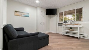 A room with a dark grey sofa, a white door, a mounted TV, a window with blinds, and a white cabinet with a radio and other items. - King Jacuzzi Suite | The Beachside Co. #9 (Sylvan Beach)