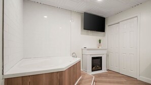 A modern bathroom with a freestanding bathtub, a white tiled wall, a fireplace, and a mounted TV. - King Jacuzzi Suite | The Beachside Co. #9 (Sylvan Beach)