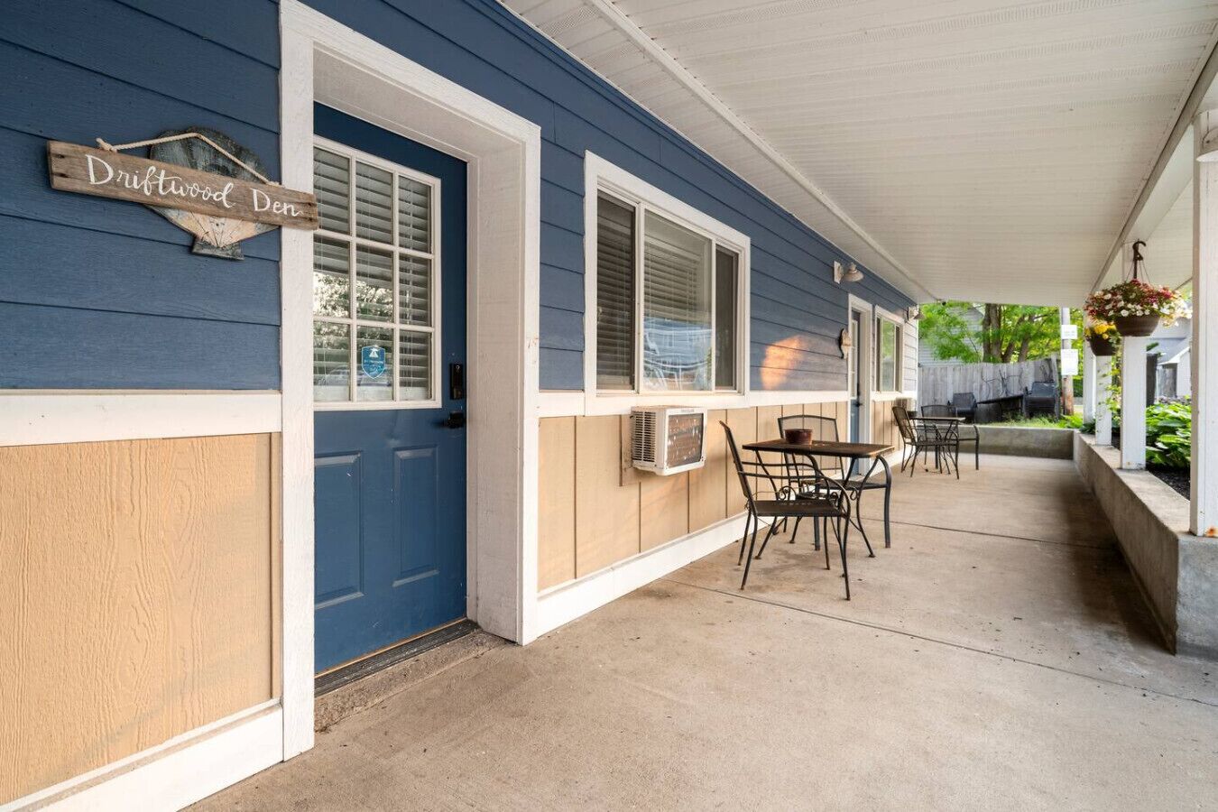 Un portico coperto con una porta blu, un cartello con scritto "Driftwood Den" e posti a sedere all'aperto.
