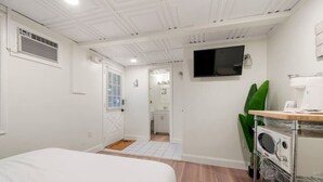 A hotel room with a bed, a TV mounted on the wall, a kitchenette with a coffee maker, and a bathroom visible through an open door. - Cozy Beach Studio | The Beachside Co. #6 (Sylvan Beach)