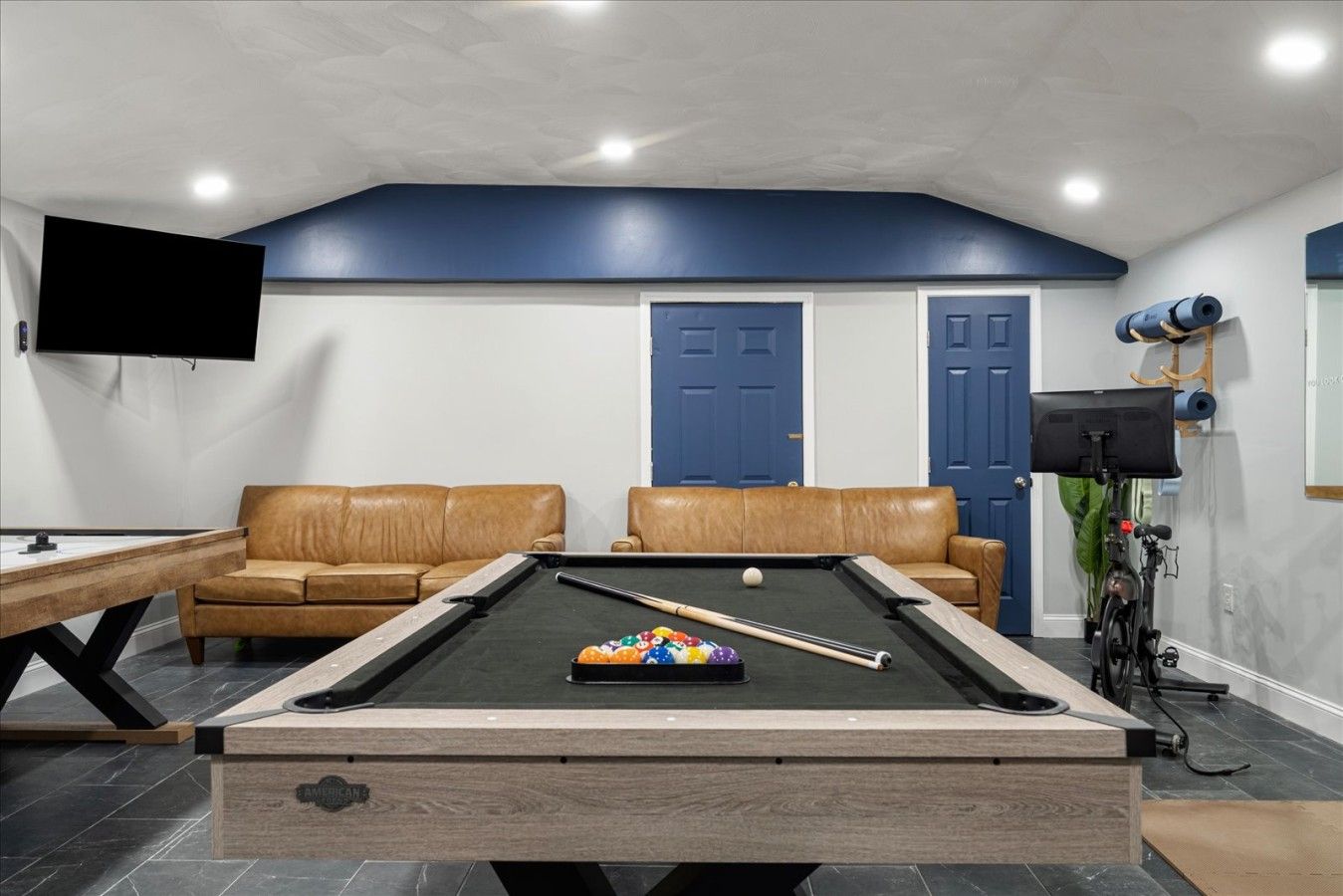 A pool table with a cue stick and balls, two leather sofas, a television mounted on the wall, and a door with a blue frame.