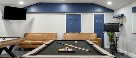 Una mesa de billar con taco y bolas, dos sofás de cuero, un televisor montado en la pared y una puerta con marco azul.