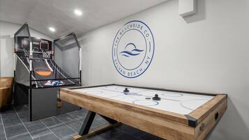 A pool table with a logo on the wall, a foosball table, and a basketball hoop in a room with a tiled floor.