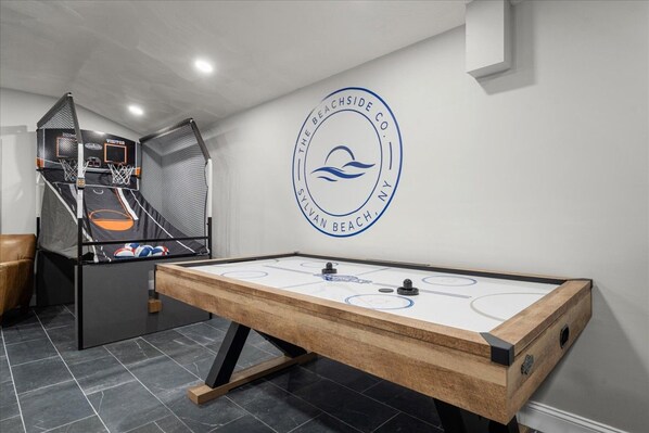 A pool table with a logo on the wall, a foosball table, and a basketball hoop in a room with a tiled floor.
