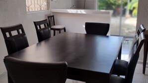 A dining area with a dark wooden table and chairs, a window with decorative bars, and a view of greenery outside.