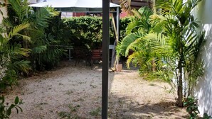 A courtyard with a gravel ground, surrounded by various plants and trees, and a white building in the background.
