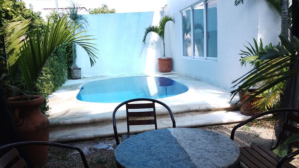 A small outdoor pool area with a stone table and chairs, surrounded by potted plants and a white building with windows.