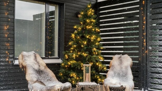 Een sneeuwbedekte veranda met twee stoelen bekleed met bont, een kerstboom met lichtjes en een lantaarn.