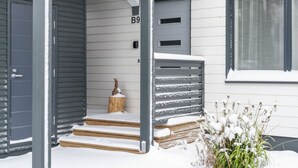Een modern huis met een grijze gevel, een houten trap die naar een deur met de aanduiding 'B9' leidt, en een klein houten paaltje met een sneeuwbedekte bovenkant.