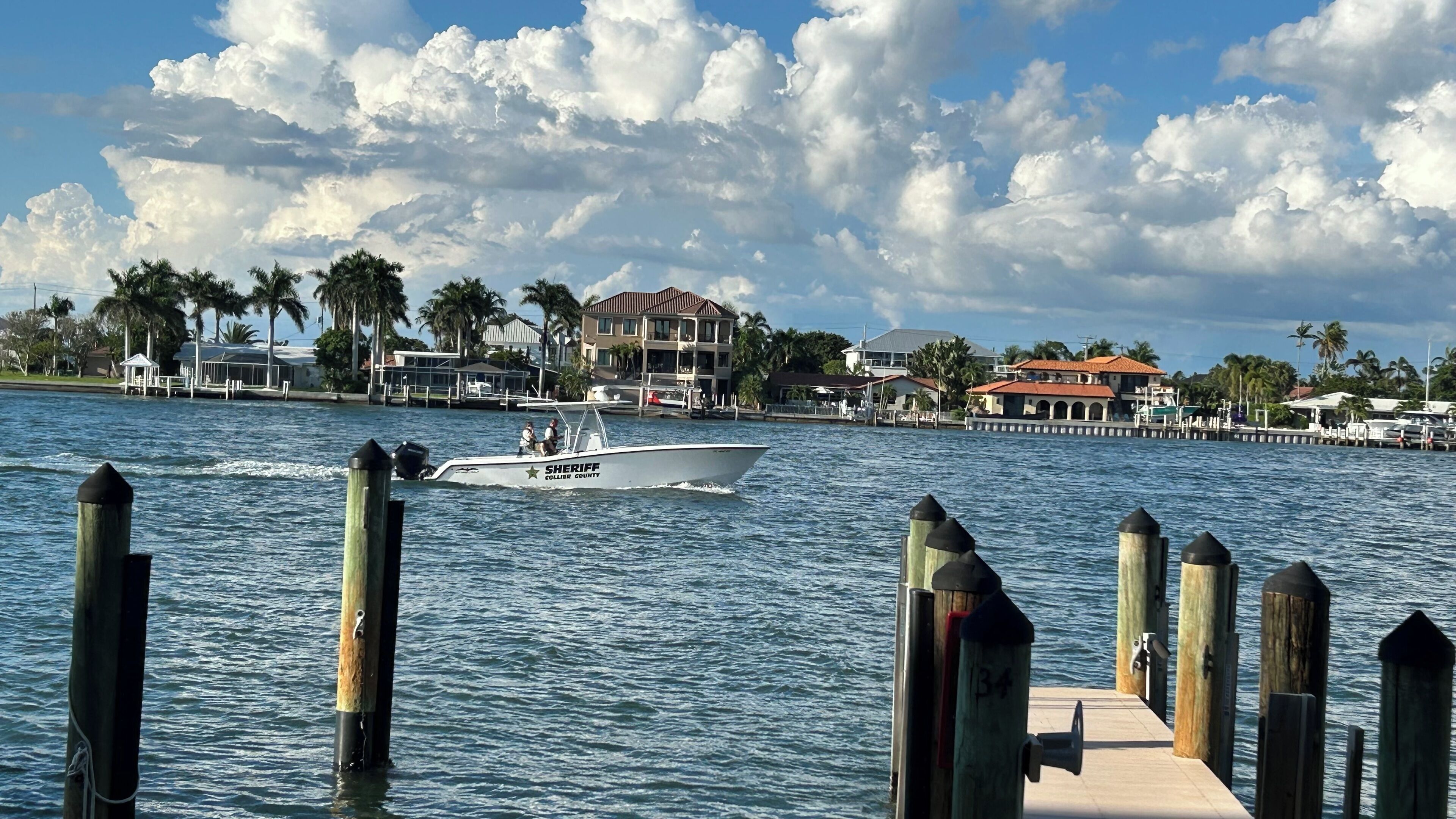 A waterfront scene with a boat named 'Sheriff' on the water, a dock with wooden posts, and buildings with palm trees in the background.