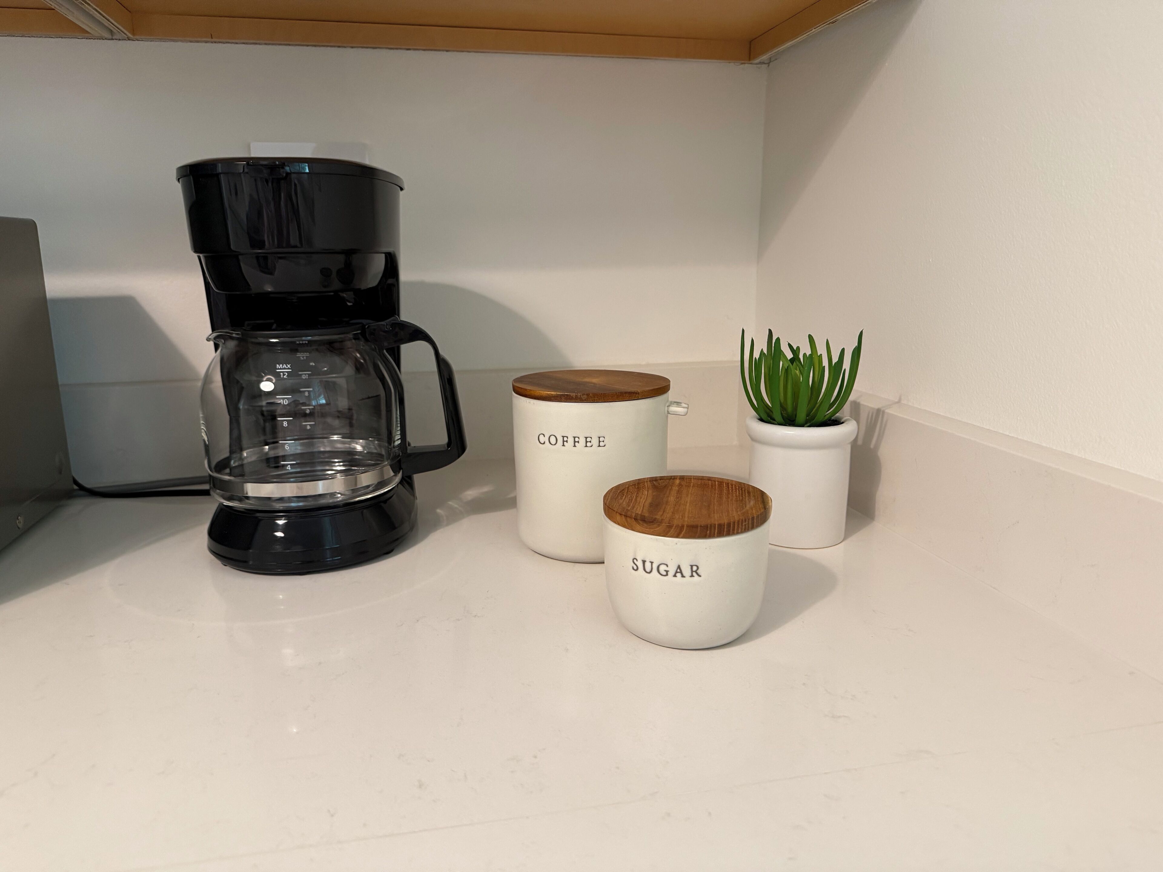Un comptoir de cuisine avec une machine à café noire, deux tasses blanches portant les inscriptions « CAFÉ » et « SUCRE », et une petite plante en pot.