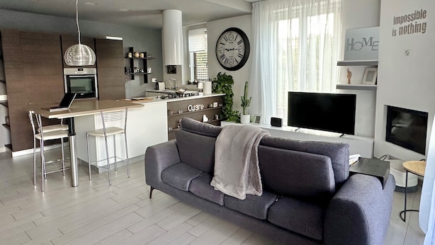 A modern living room with a grey sofa, a dining area with a kitchen in the background, a large clock, and a motivational quote on the wall. - Charming and art house in the middle of Como, Milan and lugano. (Fino Mornasco)