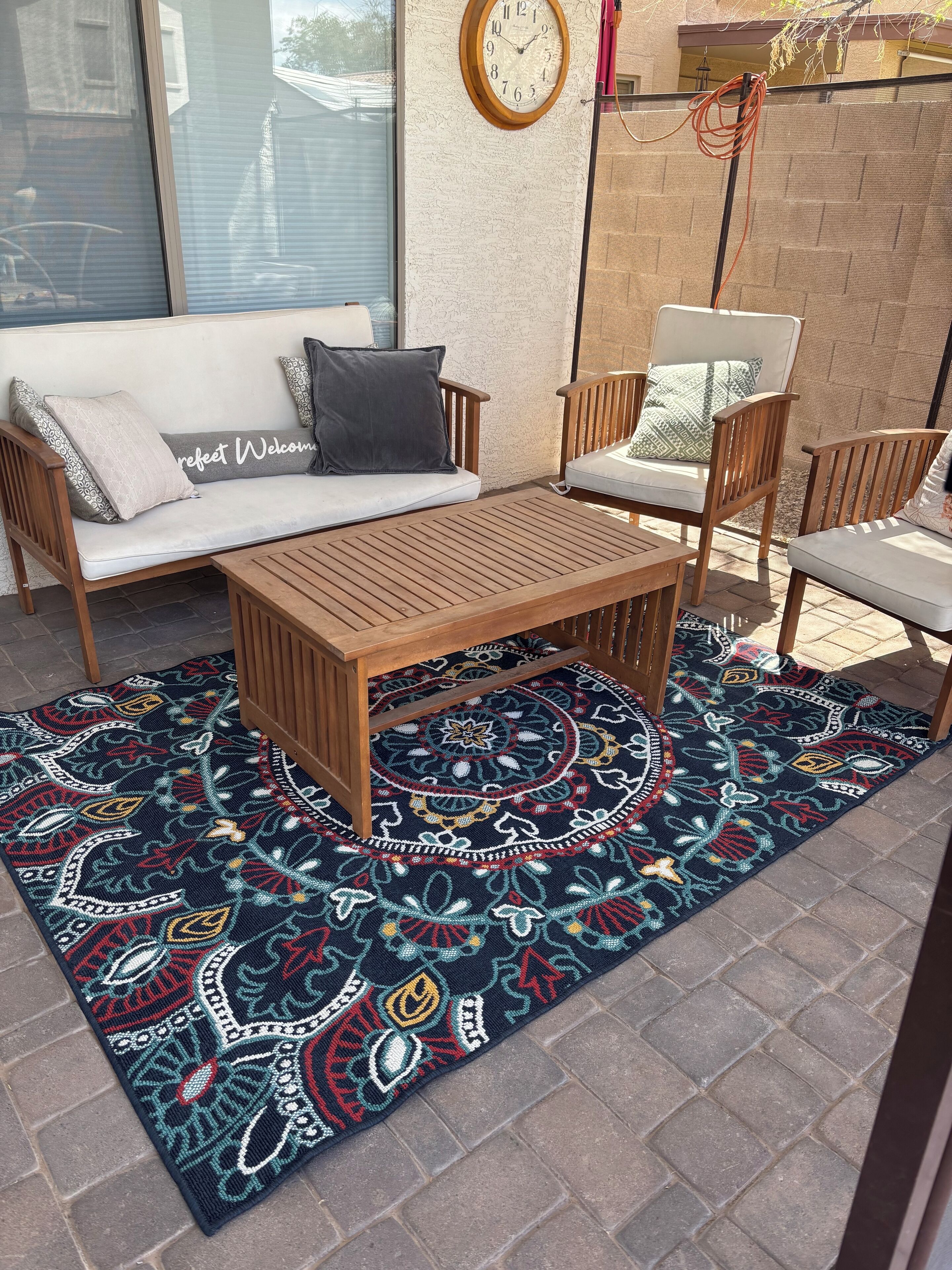 A patio area with a wooden table and chairs, a patterned rug, and a sofa with a 'Welcome' pillow.