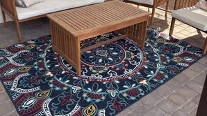 Un patio con una mesa y sillas de madera, un tapete estampado y un sofá con un cojín que dice 'Welcome'.