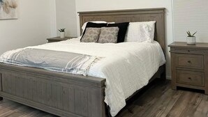 Un dormitorio con cama de madera, ropa de cama blanca, ventilador de techo y mesita de noche con una planta.