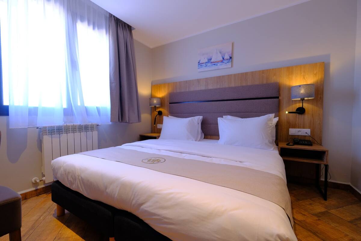 A hotel room with a large bed, two bedside tables, a headboard, a painting, and a window with curtains.