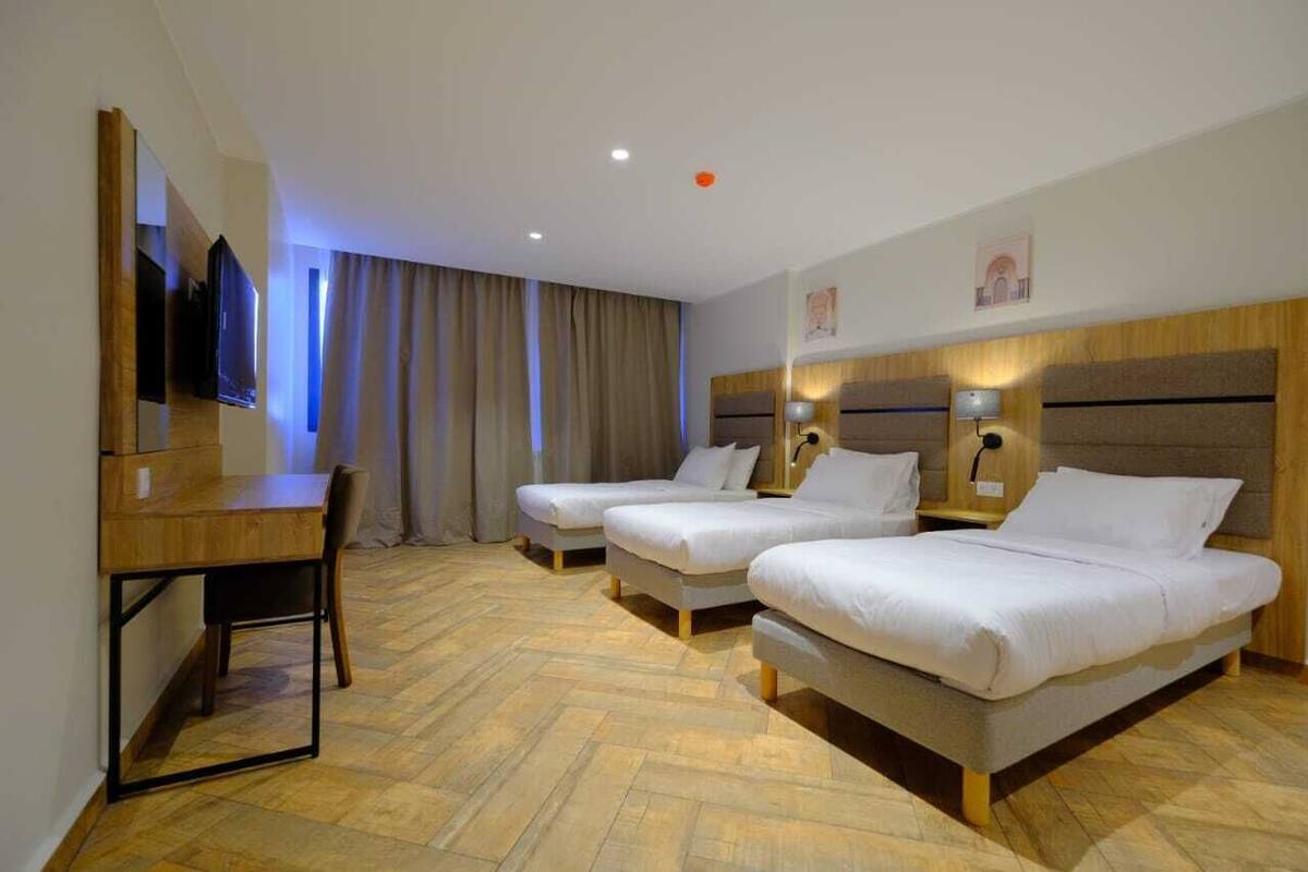 A hotel room with two single beds, a desk with a chair, a flat-screen TV mounted on the wall, and a large window with curtains.
