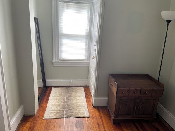 A hallway with wooden flooring, a white door, a window with blinds, a wooden cabinet, and a floor lamp.