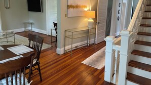 Un comedor con una mesa y sillas de madera, un televisor montado en la pared y una escalera con barandillas blancas.