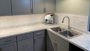 Una cocina con gabinetes grises, un salpicadero de azulejos blancos, una cafetera y un lavamanos de acero inoxidable.