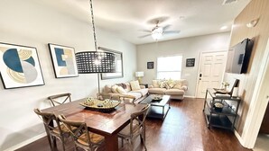 A spacious living room with a dining area, a ceiling fan, and a television mounted on the wall. - Comfy Villa 3BR Near Disney & Parks (Kissimmee)