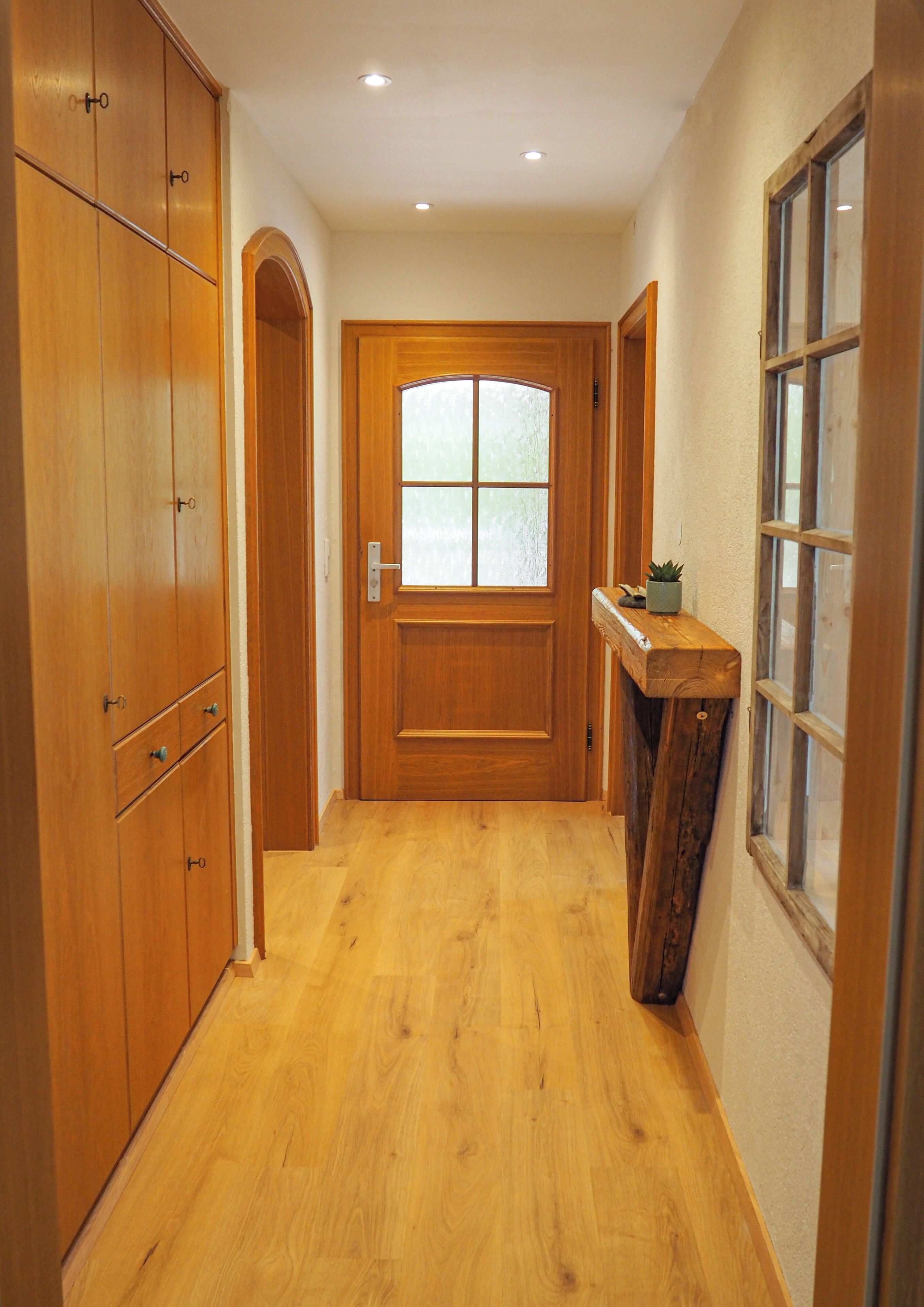 Ein Korridor mit Holzboden, einer Holztür mit Glasfenstern und einer Holzbank mit einer Pflanze.