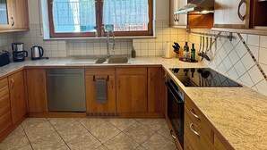 Cozinha com armários de madeira, lava-louças de aço inoxidável, janela com cortinas e painel de azulejos.
