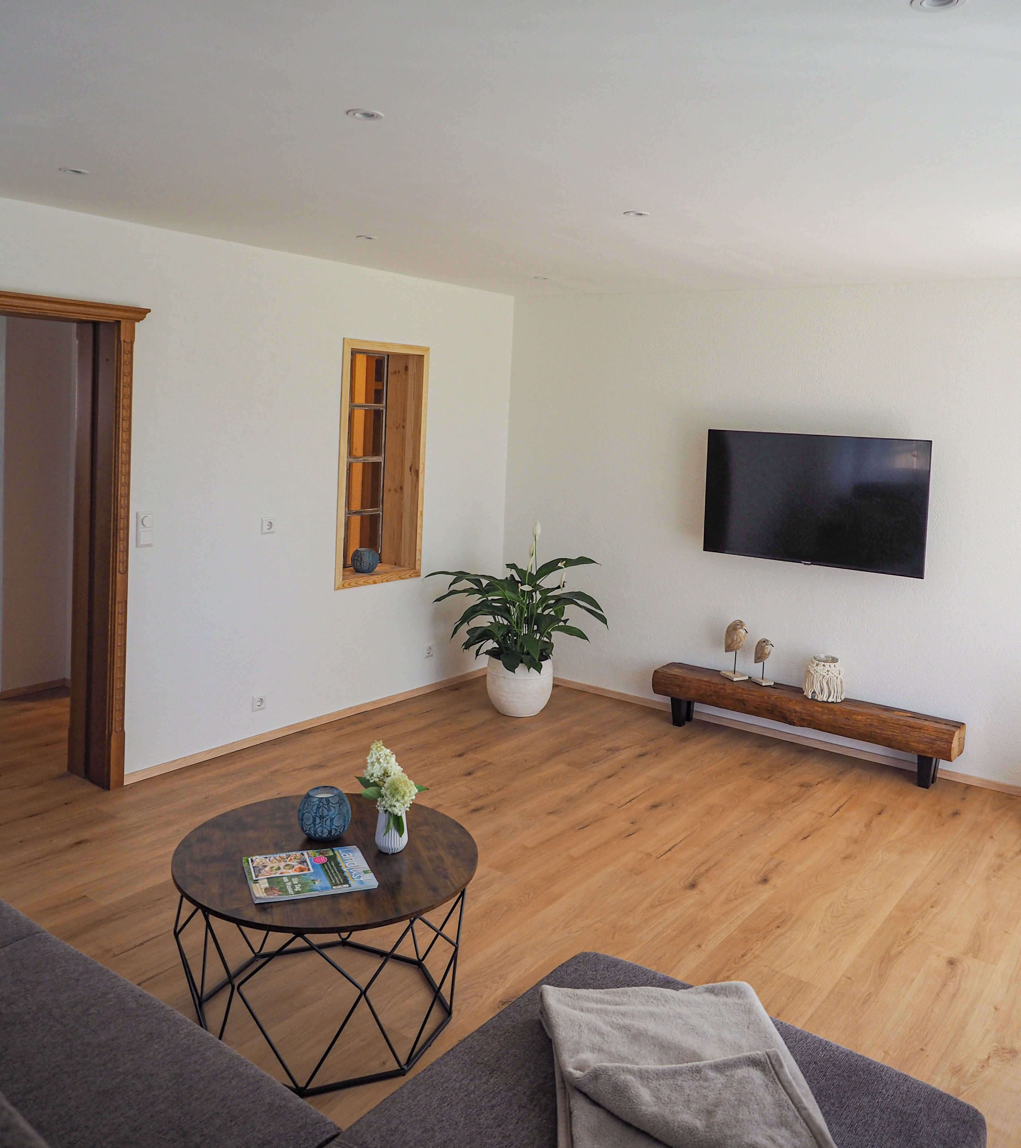 A modern living room with a wooden floor, a round coffee table with magazines, a grey sofa, a wooden bench with decorative items, a large potted plant, and a flat-screen TV mounted on the wall.