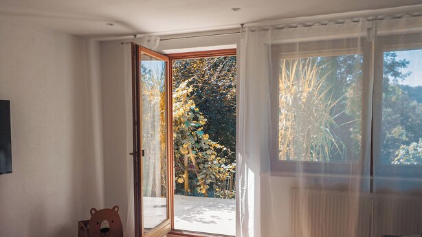 Um quarto com piso de madeira, um banco, uma mesa com um vaso decorativo e vista para um jardim através de uma porta de vidro.