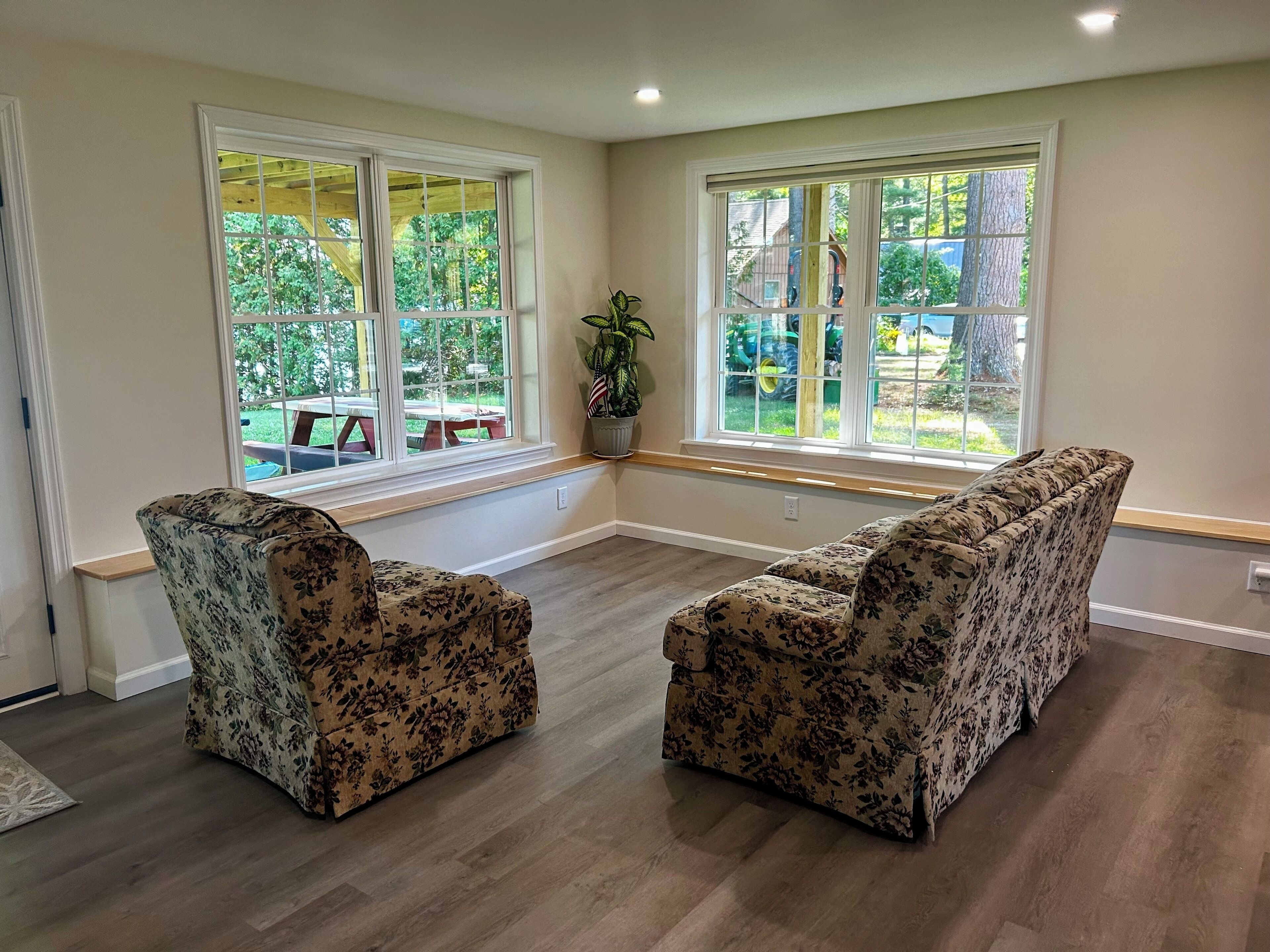 Ein geräumiges Zimmer mit zwei floralen Sofas, großen Fenstern mit Blick ins Freie und Holzboden.