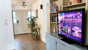 Una sala de estar con un televisor de pantalla plana sobre un mueble bajo, una estantería con diversos objetos, una planta en maceta y una puerta al fondo.